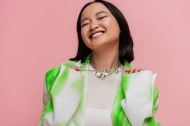 pleased asian woman in green and white clothes touching silver necklaces isolated on pink clipart