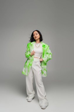 full length of asian woman in trendy oversize outfit posing with hand on hip on grey background