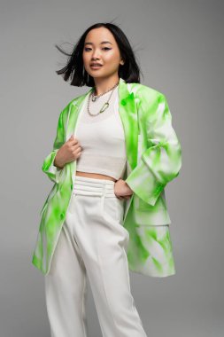 trendy asian woman in green and white blazer standing with hand on hip isolated on grey