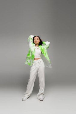 full length of brunette asian woman in white pants and trendy jacket posing with hands behind head on grey background