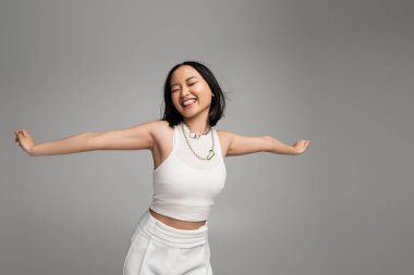 young and happy asian woman in white tank top sticking out tongue while standing with outstretched hands isolated on grey