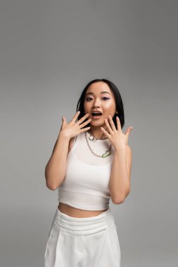 amazed asian woman in white tank top looking at camera while holding hands near face isolated on grey