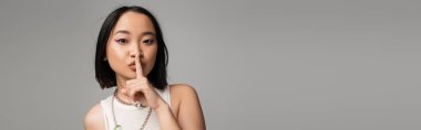 young asian woman with makeup and blue eyeliner showing hush sign isolated on grey, banner