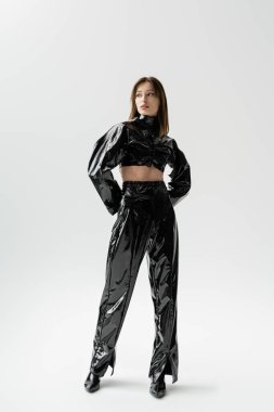 full length of stylish woman in black latex outfit and high heels standing on grey 