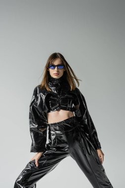 stylish woman in black latex outfit and sunglasses posing isolated on grey