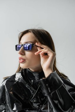 young woman in black latex jacket adjusting trendy sunglasses isolated on grey
