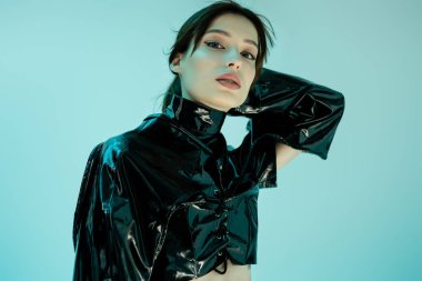 young woman in black latex clothing looking at camera isolated on blue 