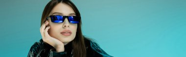 young woman in black latex clothing and sunglasses isolated on blue, banner 