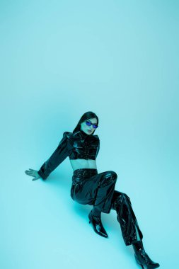 full length of young woman in sexy latex clothing and high heels sitting on blue background 