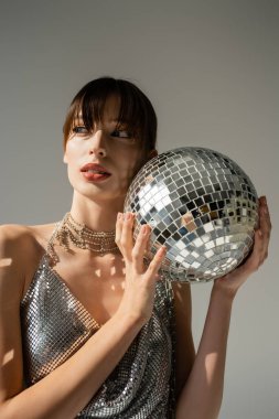 stylish woman in shiny top holding disco ball isolated on grey
