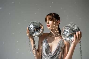 young woman in trendy and shiny top holding chain with disco balls on grey  clipart