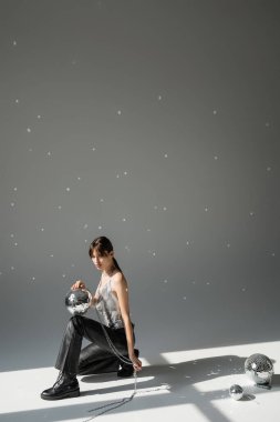 full length of brunette woman in trendy top and leather trousers holding chain with shiny disco ball on grey 