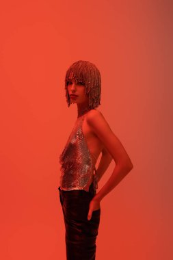 Stylish young woman in jewelry headwear posing and looking at camera isolated on red 