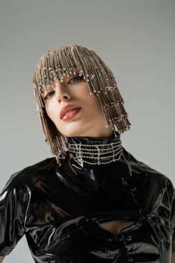 Portrait of stylish woman in latex top and jewelry headwear looking at camera isolated on grey 