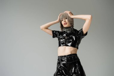 Trendy young model in latex clothes and jewelry headwear looking at camera isolated on grey 