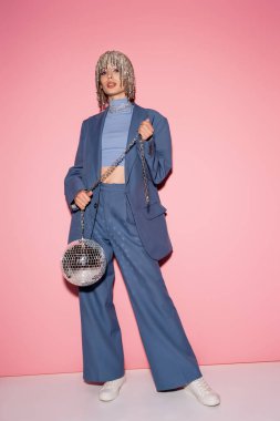 Trendy woman in suit and luxury headwear holding disco ball on pink background 