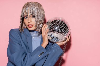 Portrait of trendy young woman in jewelry headwear holding disco ball on pink background 
