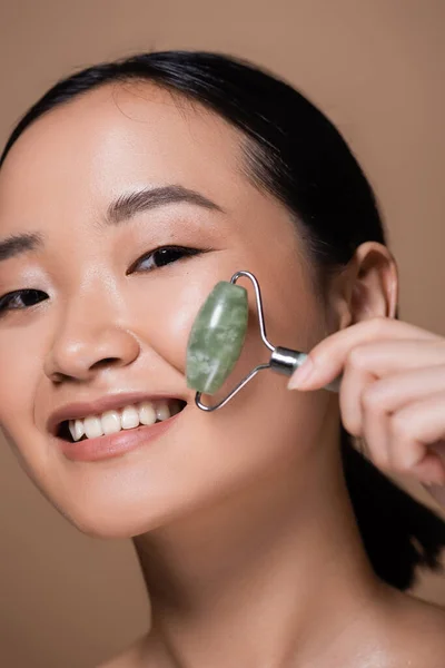 stock image Portrait of cheerful asian woman holding jade roller and looking at camera isolated on brown 