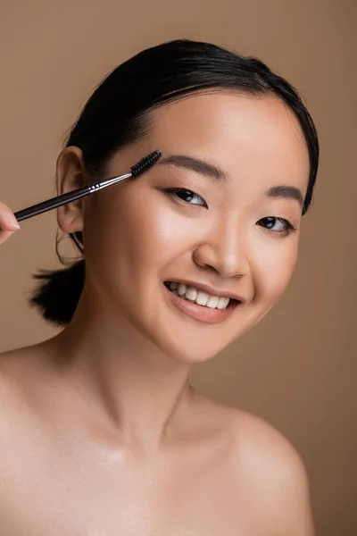 Smiling asian model holding eyebrow brush and looking at camera isolated on brown