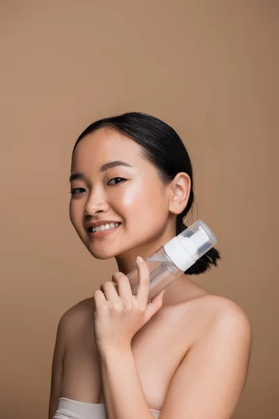 Young asian woman with naked shoulders holding face cleanser isolated on brown