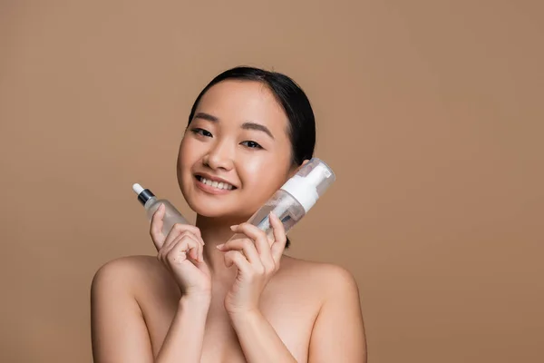 Cheerful asian model holding serum and face foam isolated on brown