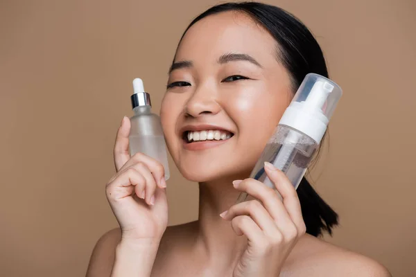 Positive asian woman with naked shoulders holding serum and face cleanser isolated on brown