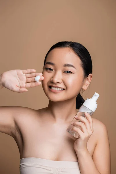 Pretty asian woman in top holding cleansing foam isolated on brown