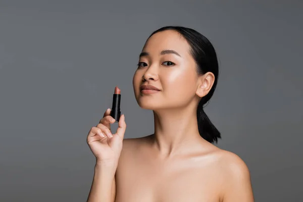 stock image Pretty asian woman with naked shoulders holding beige lipstick isolated on grey 