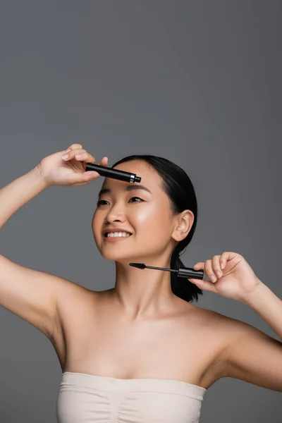 stock image cheerful asian woman in top holding mascara and eyelash brush isolated on grey