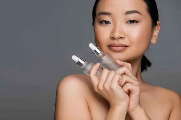 asian woman with perfect face holding bottles of cosmetic serum while looking at camera isolated on grey