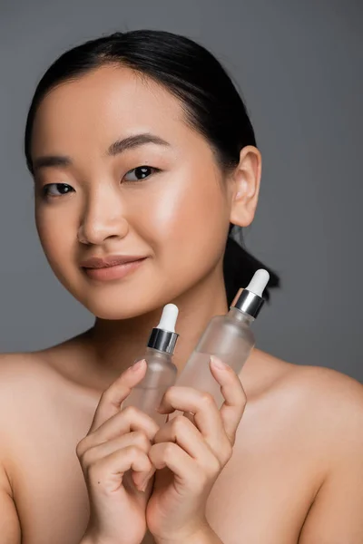 stock image pleased asian woman with naked shoulders holding bottles with cosmetic serum and looking at camera isolated on grey