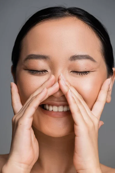 young asian woman with closed eyes and nude makeup smiling and touching face isolated on grey