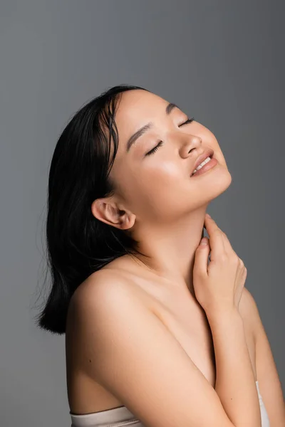 pleased asian woman with closed eyes and nude makeup touching neck isolated on grey