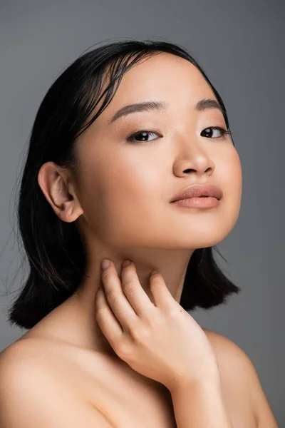 stock image portrait of asian woman with bare shoulders and natural makeup touching neck and looking at camera isolated on grey