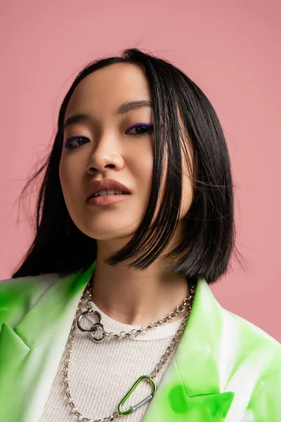 portrait of brunette asian woman in silver necklaces and makeup looking at camera isolated on pink