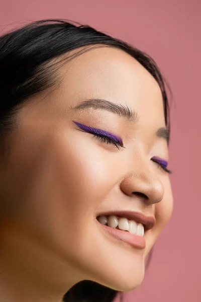 stock image close up view of young asian woman with makeup and blue eyeliner smiling with closed eyes isolated on pink