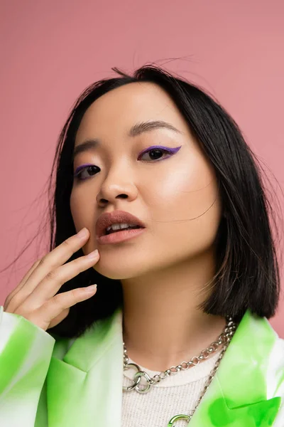 portrait of stylish asian woman in silver necklaces touching face with makeup and looking at camera isolated on pink