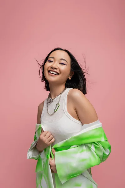 stock image young and stylish asian woman in silver necklaces and white tank top looking at camera on wind isolated on pink