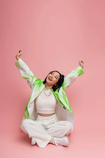 stock image joyful asian woman in stylish outfit sitting with crossed legs and stretching raised hands on pink background