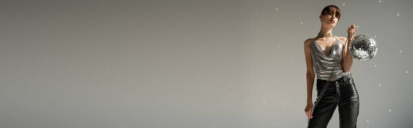 stylish woman in shiny top and leather pants holding disco ball on grey background, banner 