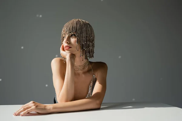 stock image young model with metallic headwear with jewelry looking away while sitting at desk on grey 