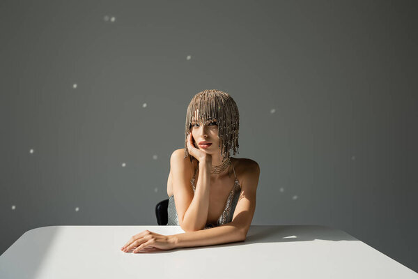 young model with metallic headwear with jewelry looking at camera while leaning on table on grey 