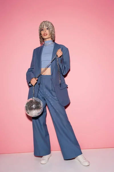 stock image Trendy woman in suit and luxury headwear holding disco ball on pink background 