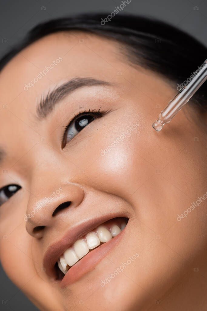 Close up view of smiling asian woman applying cosmetic serum during facial pampering isolated on grey