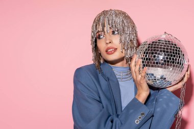 Trendy young woman in jewelry headwear holding disco ball on pink background 