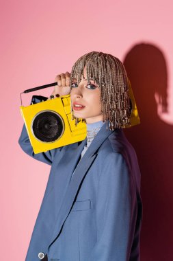 Trendy woman in jacket and jewelry headwear holding boombox on pink background 