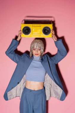 Fashionable woman in luxury headwear and suit holding boombox above head on pink background 