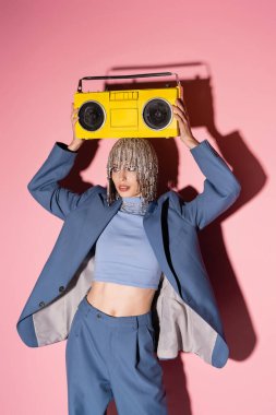 Stylish model in jewelry headwear and blazer holding boombox above head on pink background 