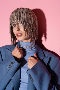 Portrait of young woman in jewelry headwear touching jacket on pink background 