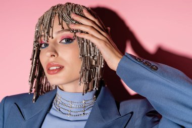 Portrait of pretty young woman with visage touching headwear with rhinestones on pink background 